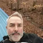 A man with a beard is sitting in a hammock in the woods.