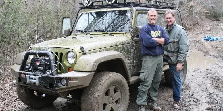 Two men are standing next to a jeep on a muddy road.