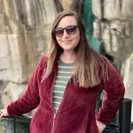 A woman wearing sunglasses and a red jacket is standing in front of a stone wall.