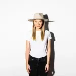 A woman wearing a hat and a white t-shirt is standing in front of a white wall.