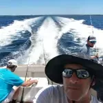 A man wearing a hat and sunglasses is taking a selfie on a boat in the ocean.