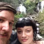 A man and a woman are posing for a picture in front of a waterfall.