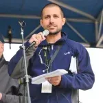 A man is holding a piece of paper and speaking into a microphone.