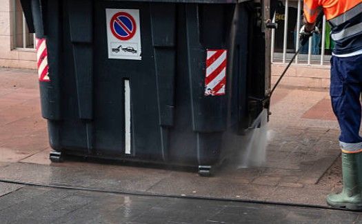 Person in work clothes power washing a large, dark dumpster on a city street.