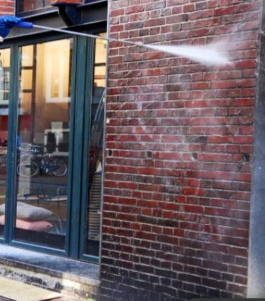 Pressure washing a red brick wall next to a glass door and window.