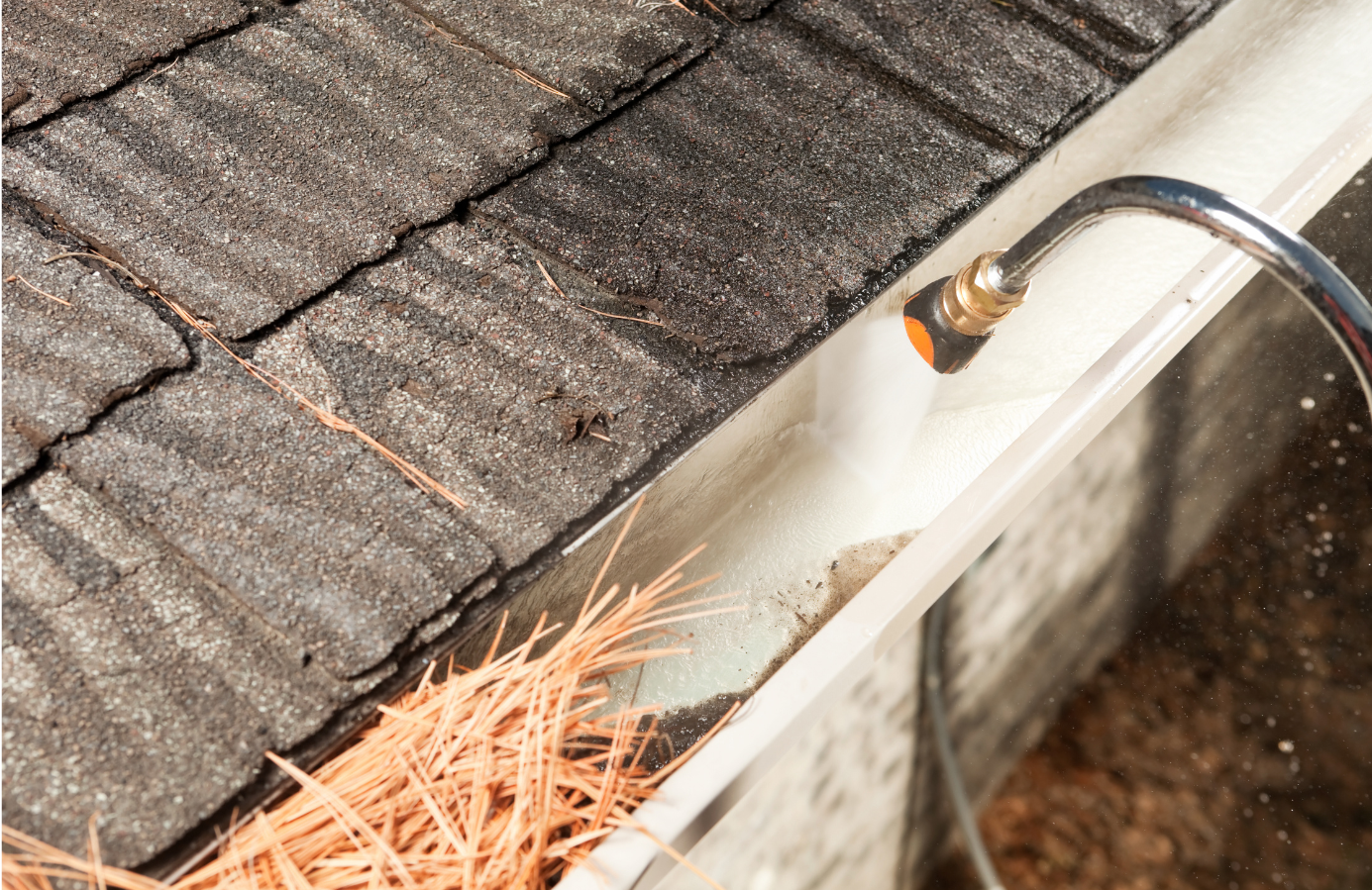 Cleaning a gutter with a pressure washer; debris and orange pine needles are visible.