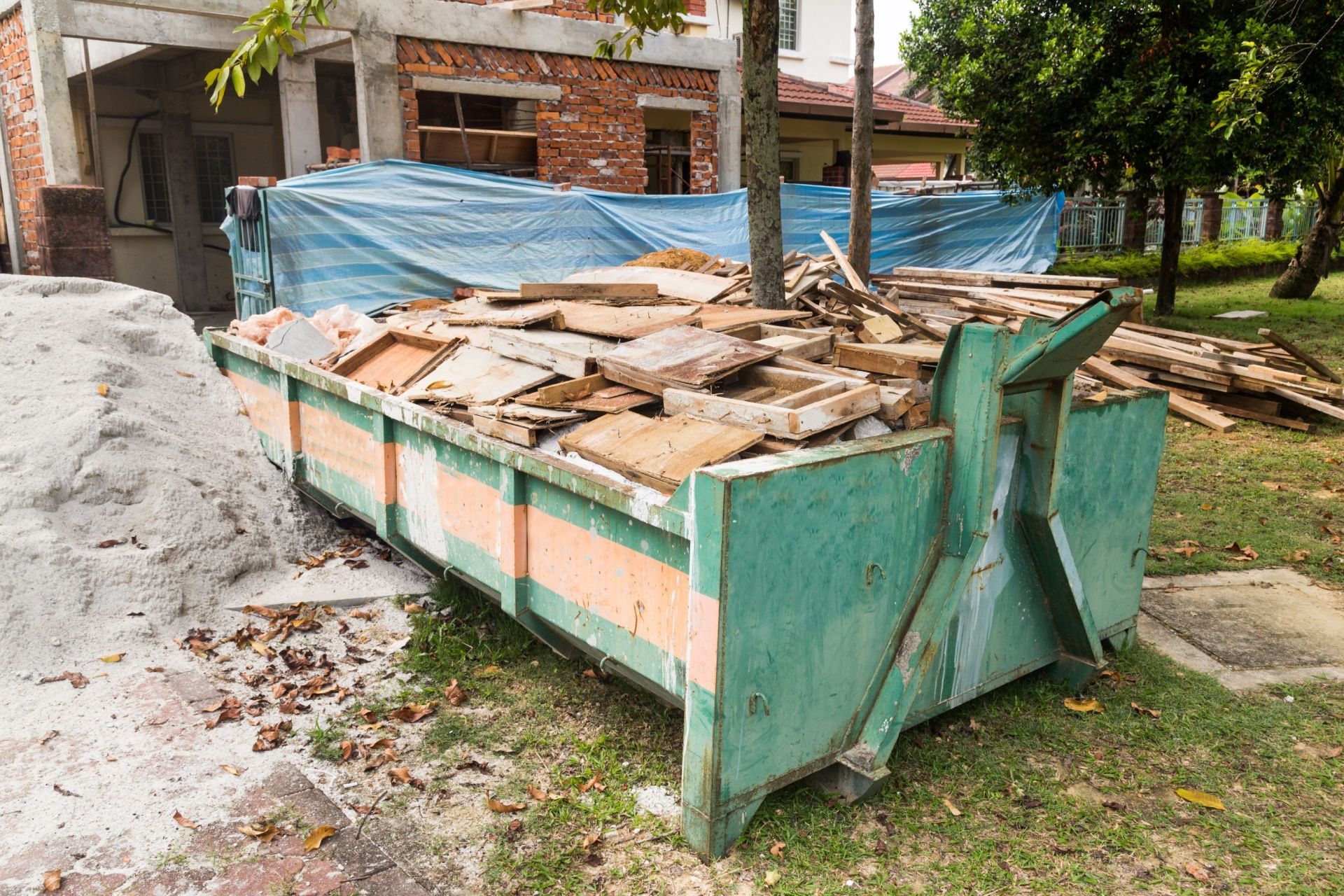 A dumpster filled with wood is sitting in the grass in front of a building under construction.