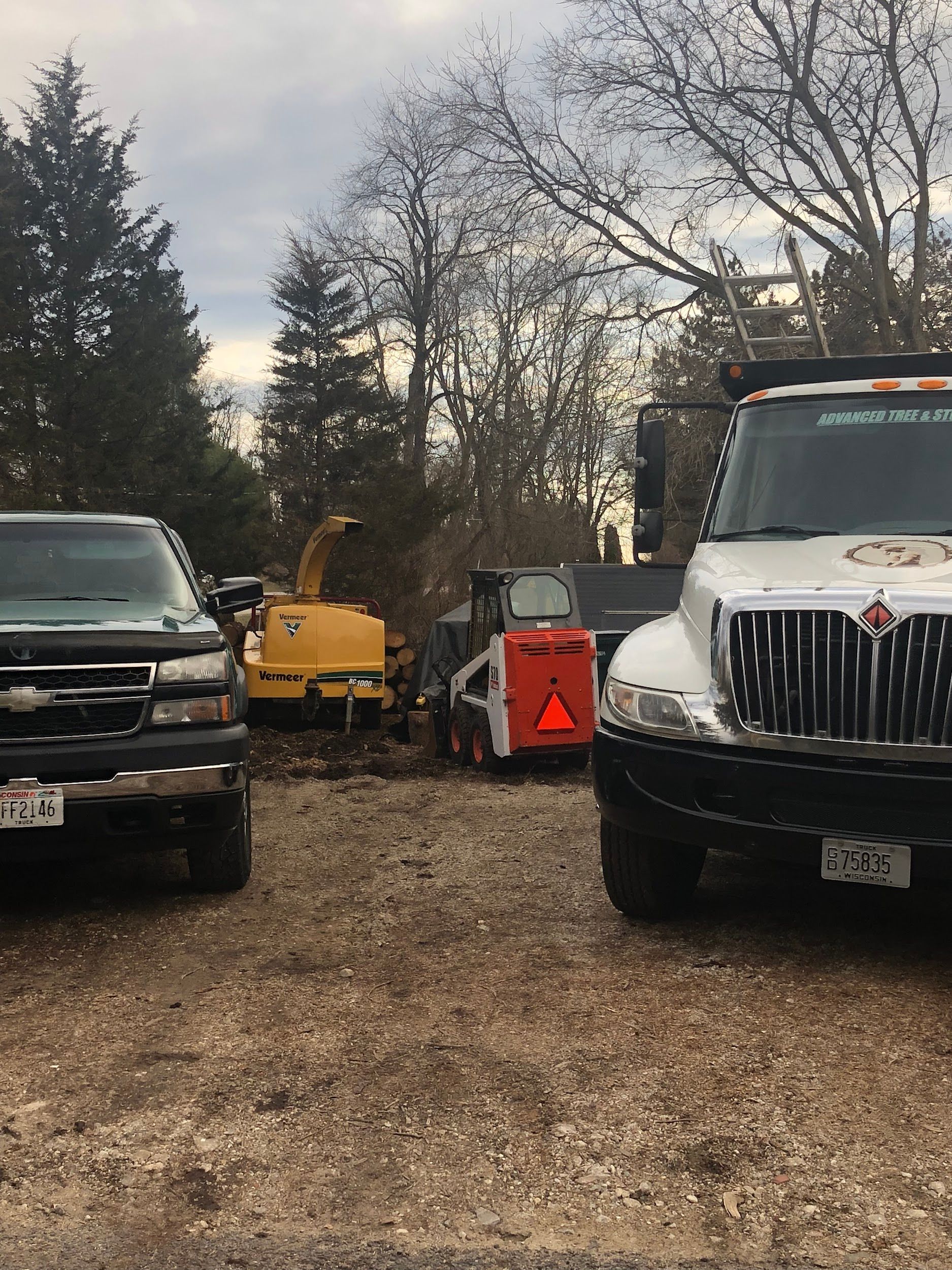 A work site with a truck, a pickup, and equipment including a chipper and a Bobcat, under a cloudy sky.