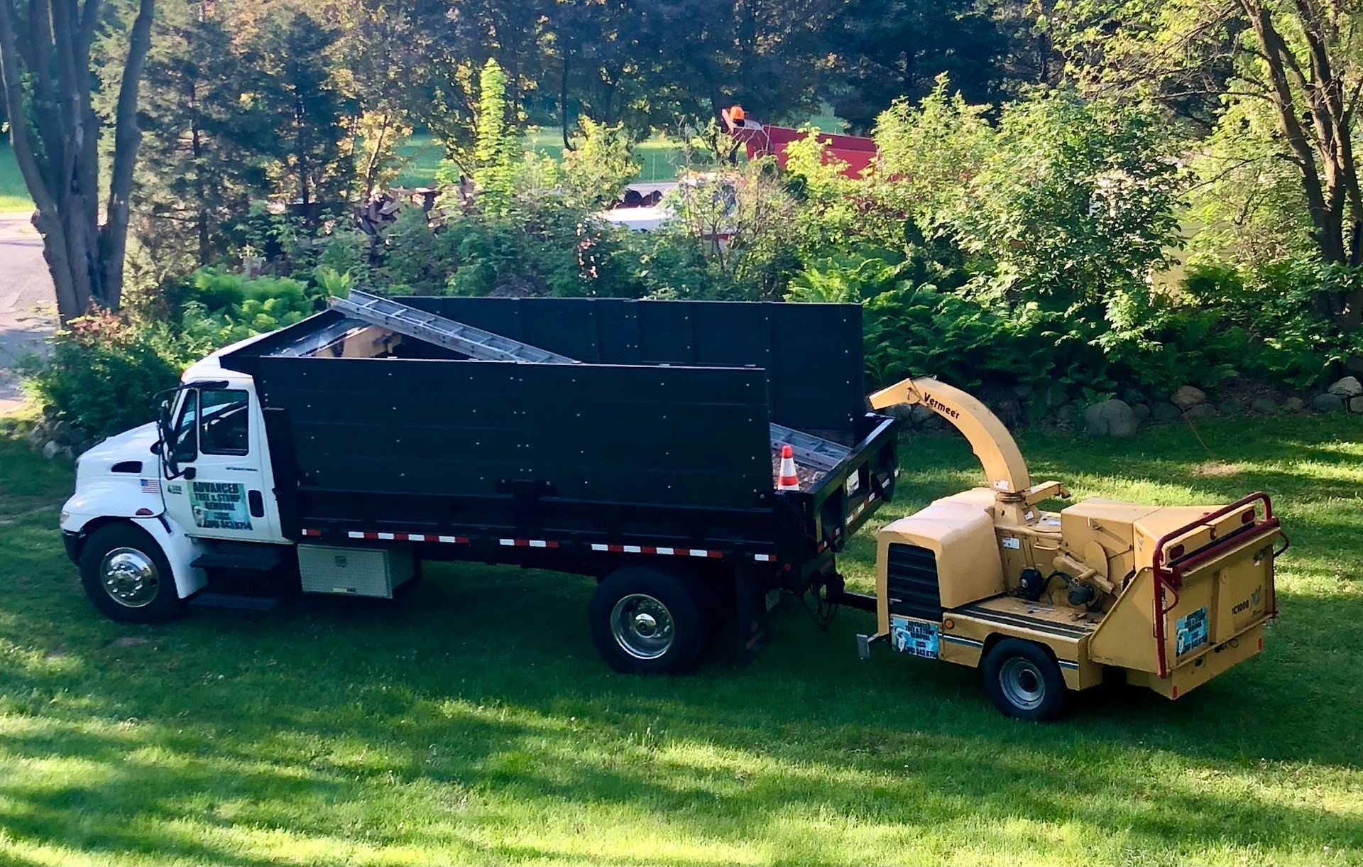 White dump truck and yellow wood chipper on grass, trees in background.