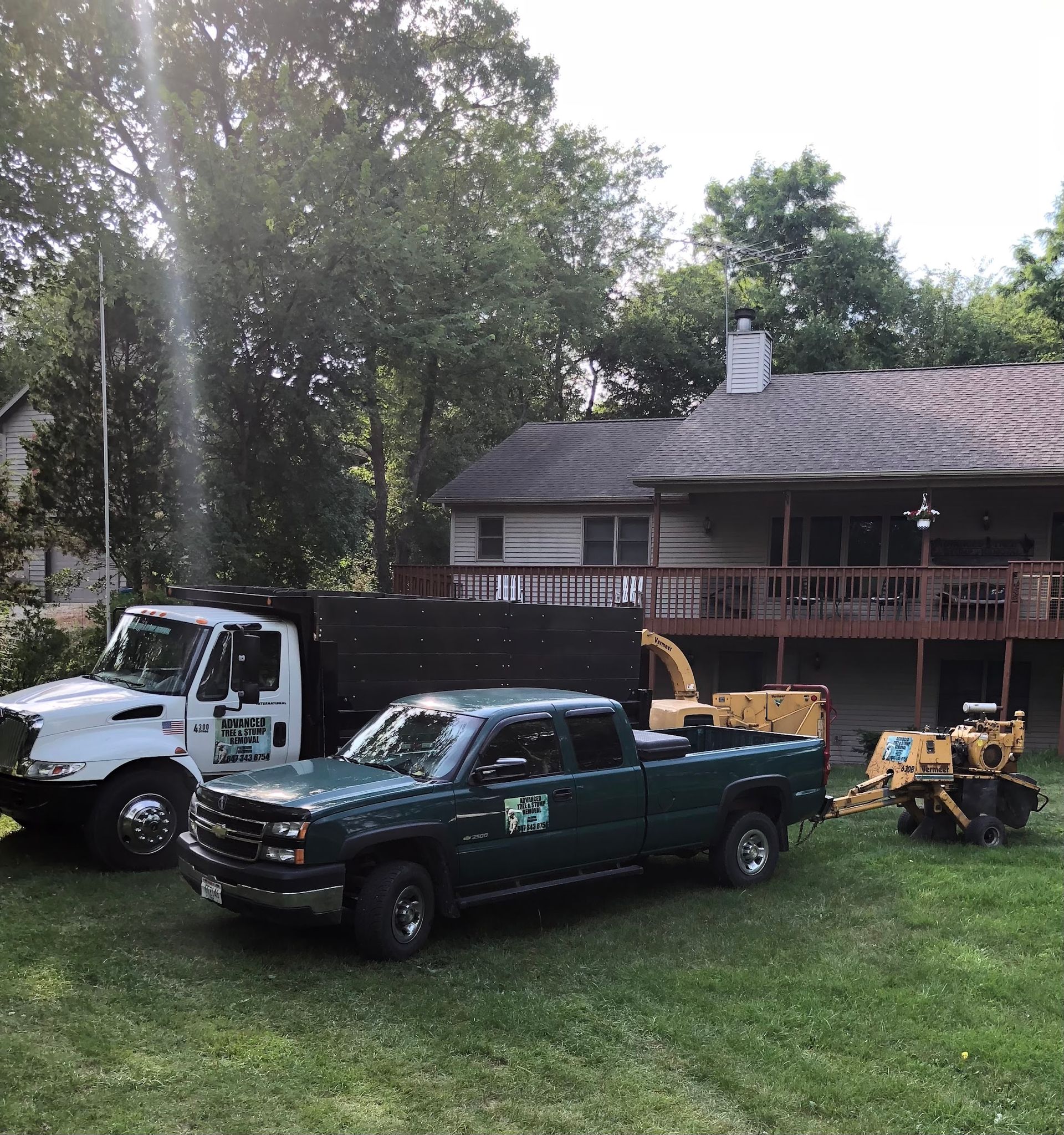 Tree service trucks parked in front of a house: dump truck, green pickup truck, and a chipper.