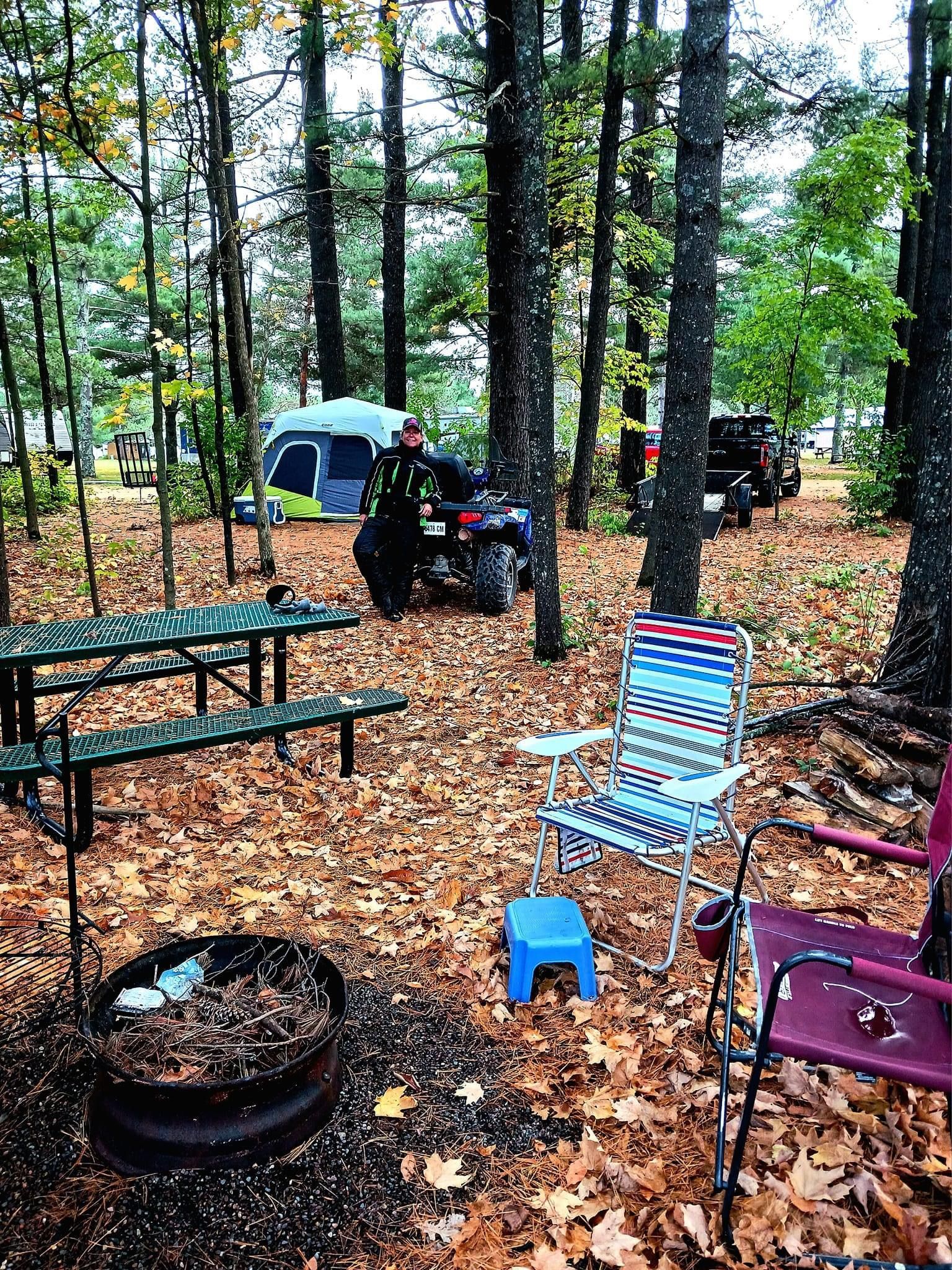A person is standing in the woods next to a picnic table and chairs.