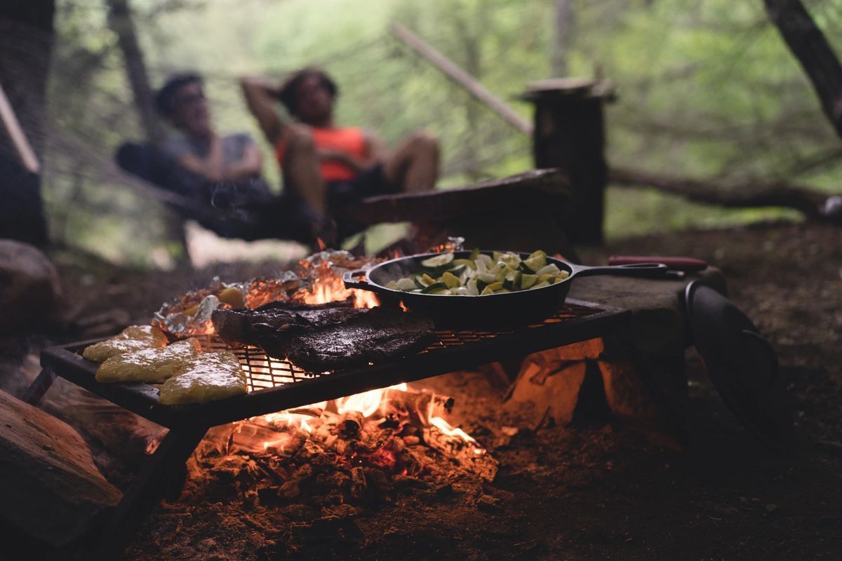A man and a woman are sitting in a hammock while cooking food over a campfire.
