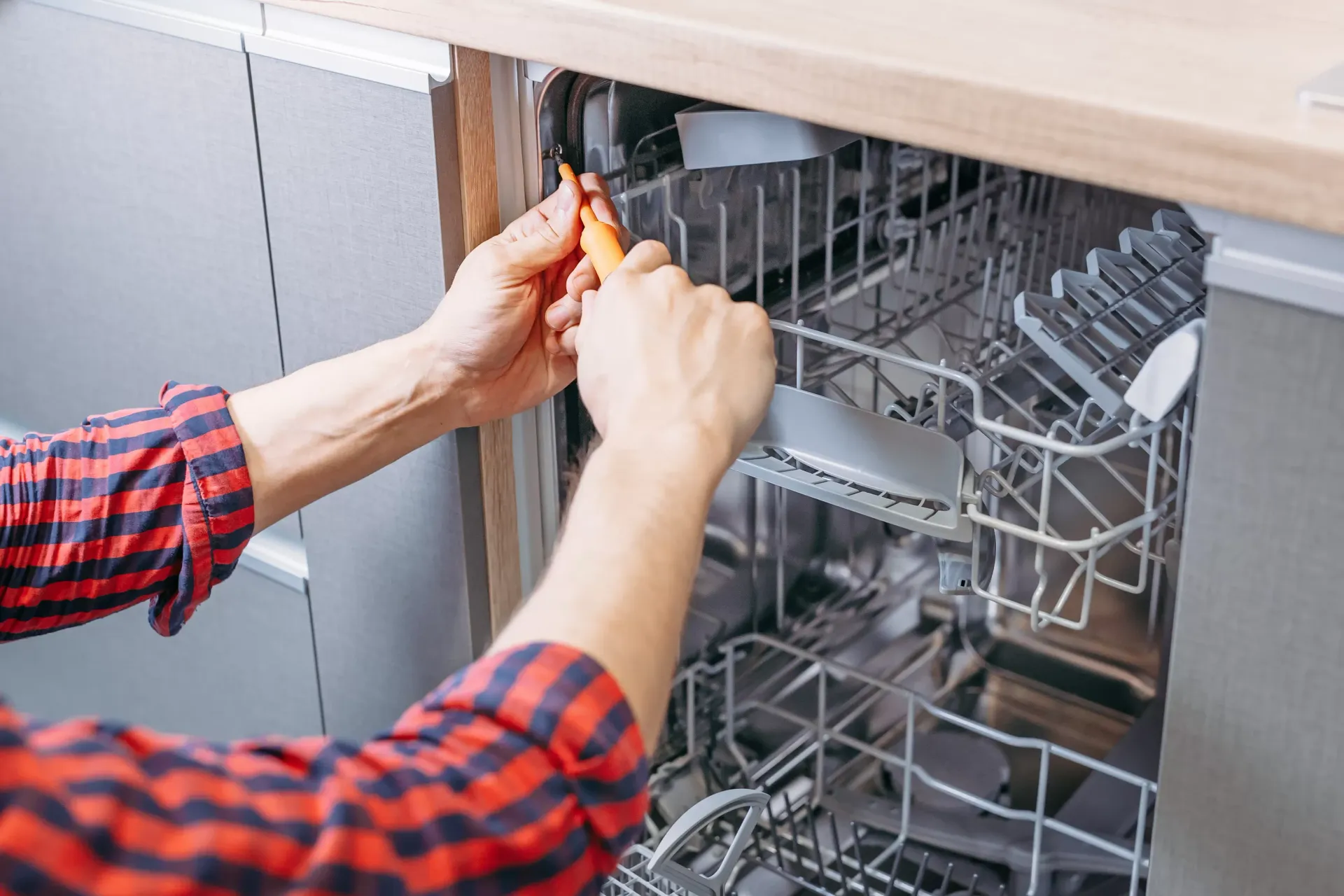 A man is fixing a dishwasher in a kitchen with a screwdriver.