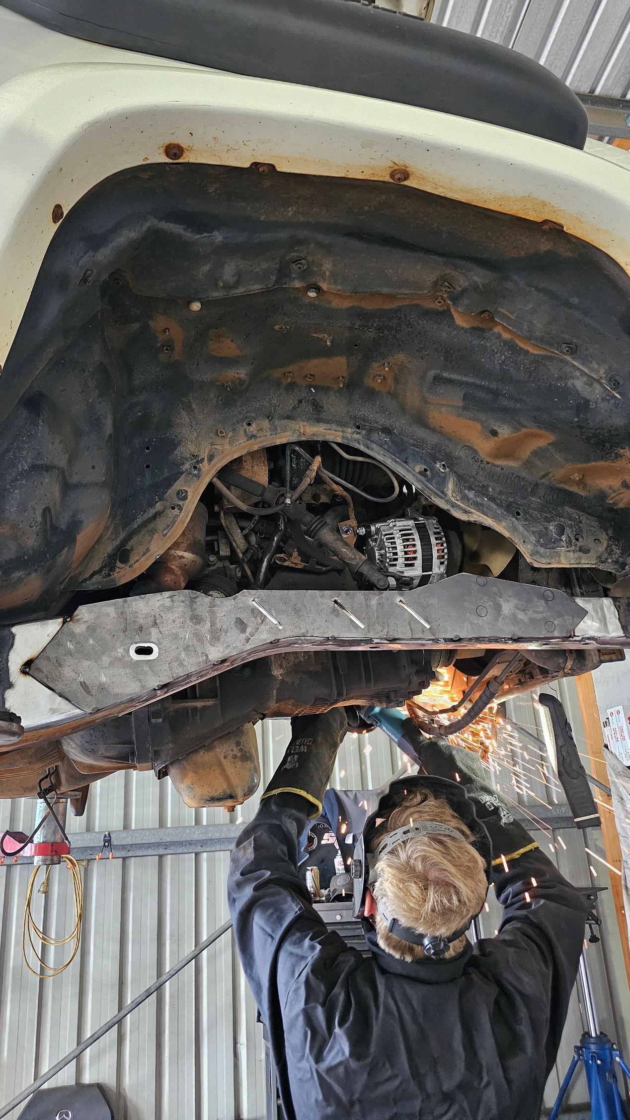 A person welds beneath a vehicle, sparks flying. Brown rust on the wheel well and frame.