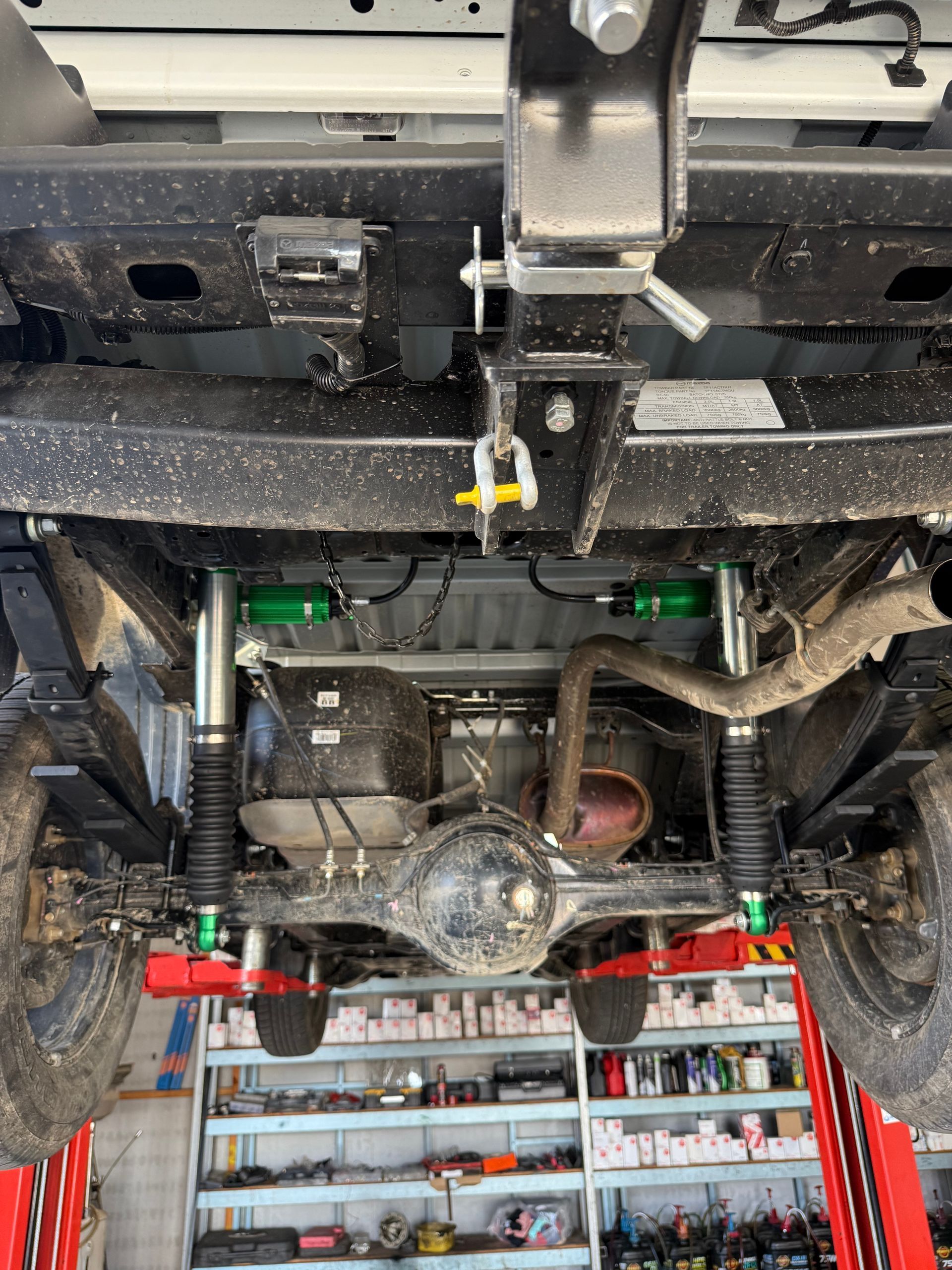 Undercarriage of a vehicle lifted on a hoist. Green suspension components, towing hitch, and rusty exhaust are visible — Track N Back 4x4 In Mareeba, QLD