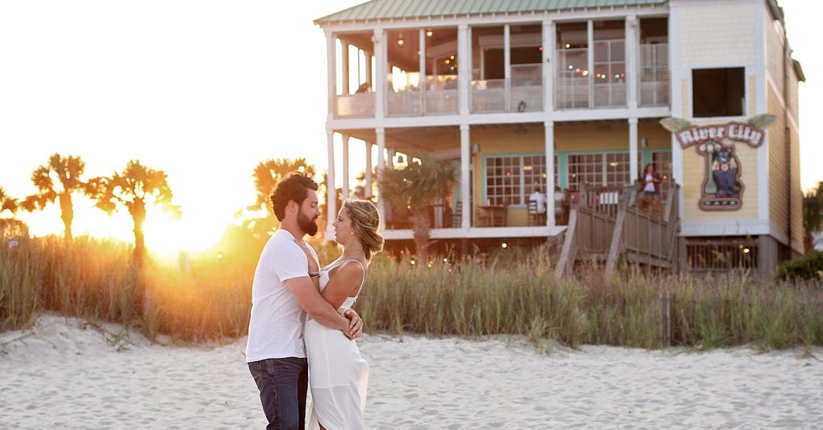 A man is holding a woman in his arms on the beach in front of a house.
