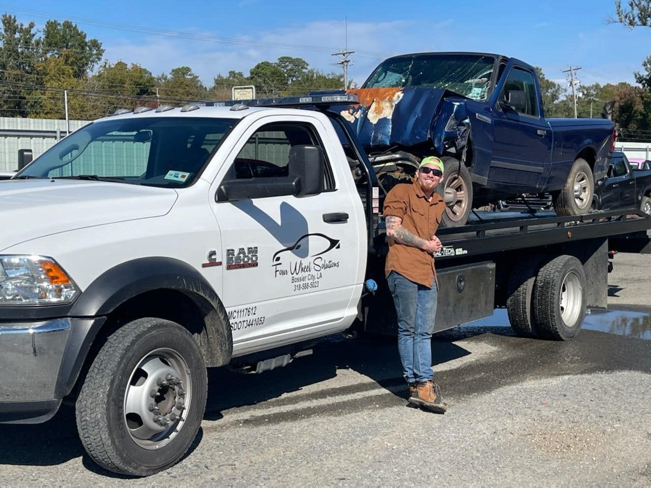 A man is standing next to a tow truck with a blue truck on the back.