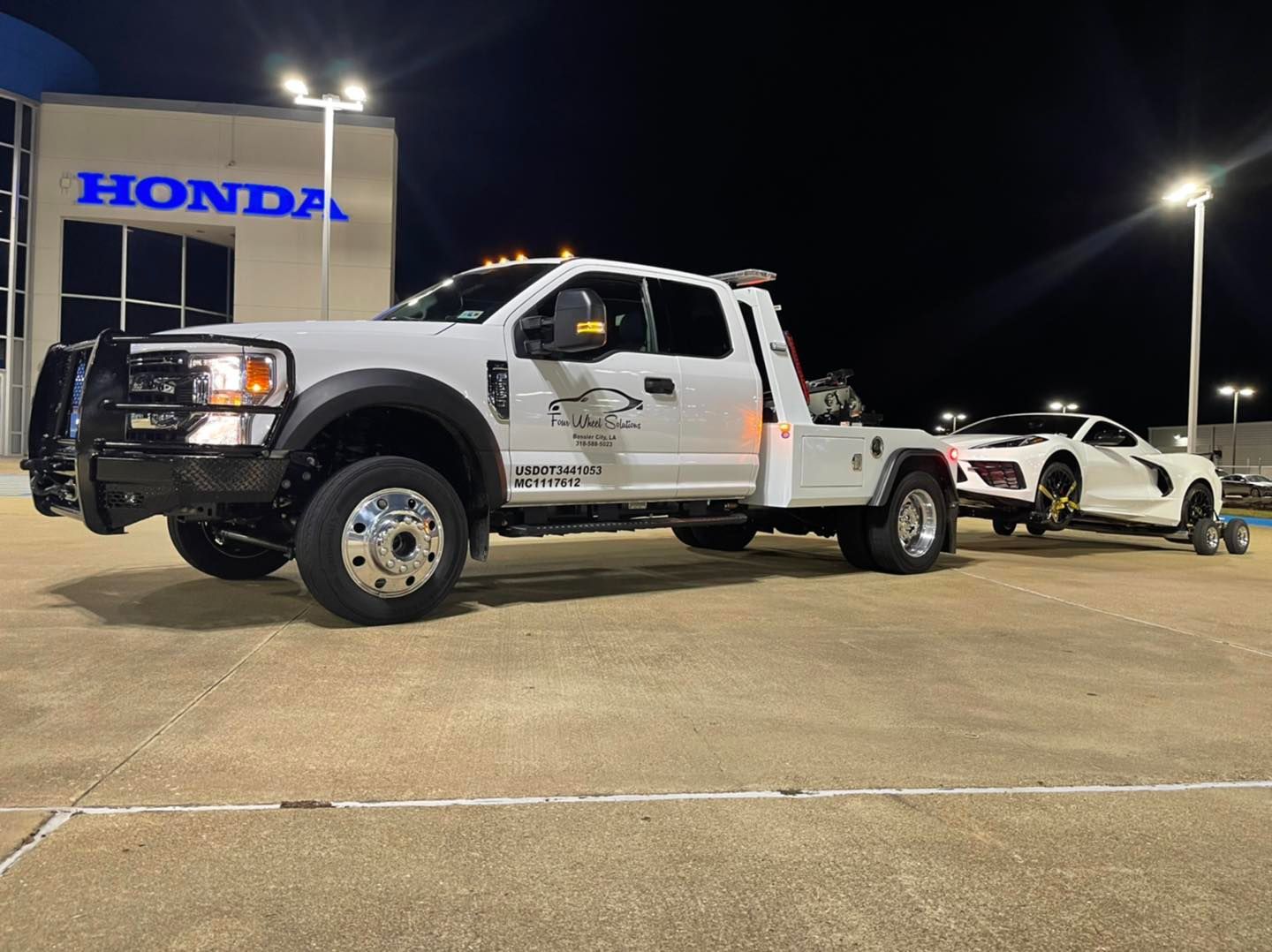 A white tow truck is towing a white car in front of a honda dealership.