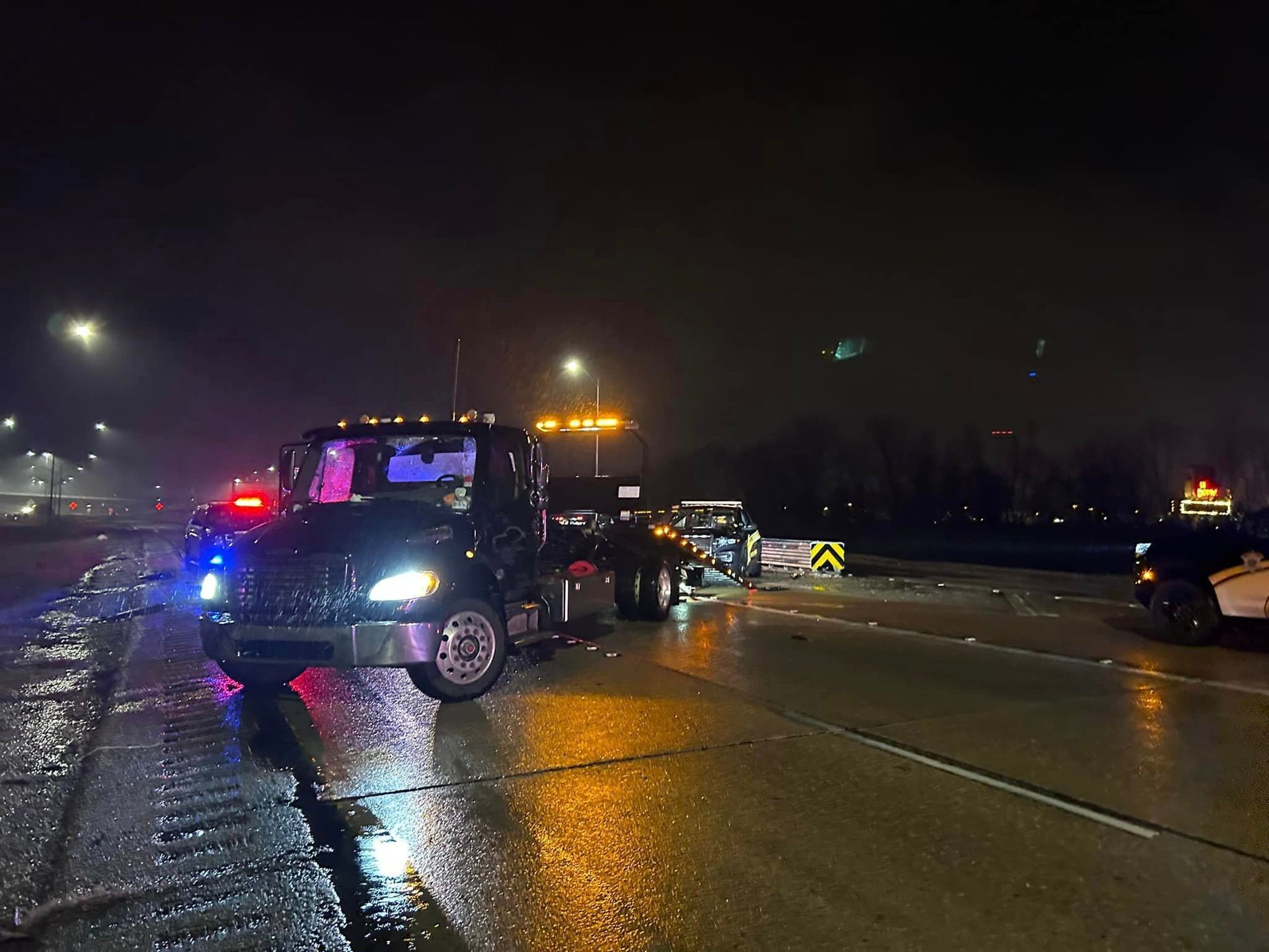 A tow truck is towing a car on a wet road at night.