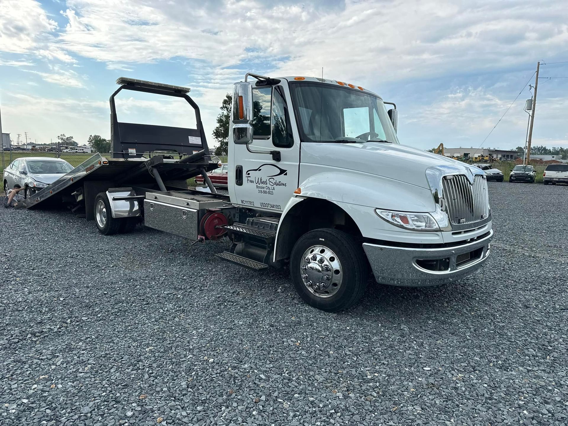 A white tow truck with a car on the back is parked in a gravel lot.