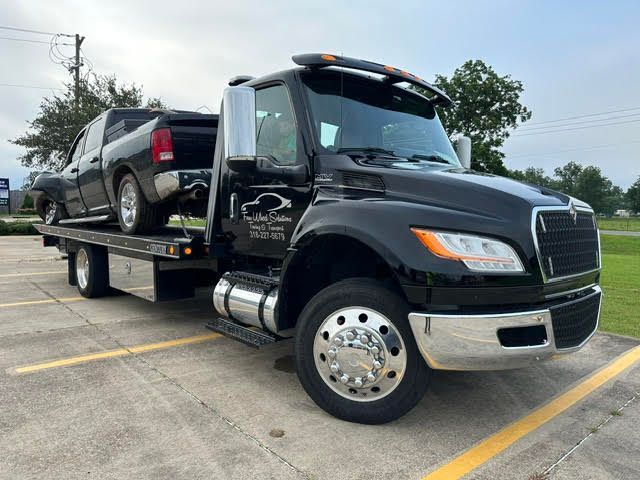 A black tow truck with a truck on the back is parked in a parking lot.