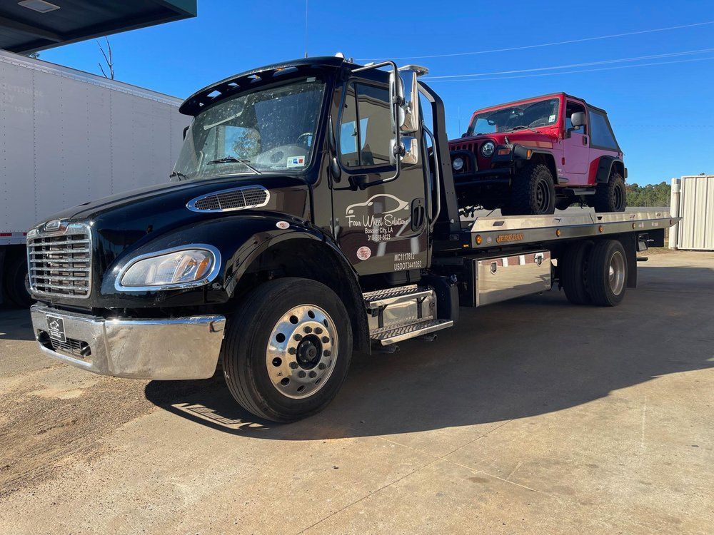 A black tow truck with a red jeep on the back is parked in a parking lot.