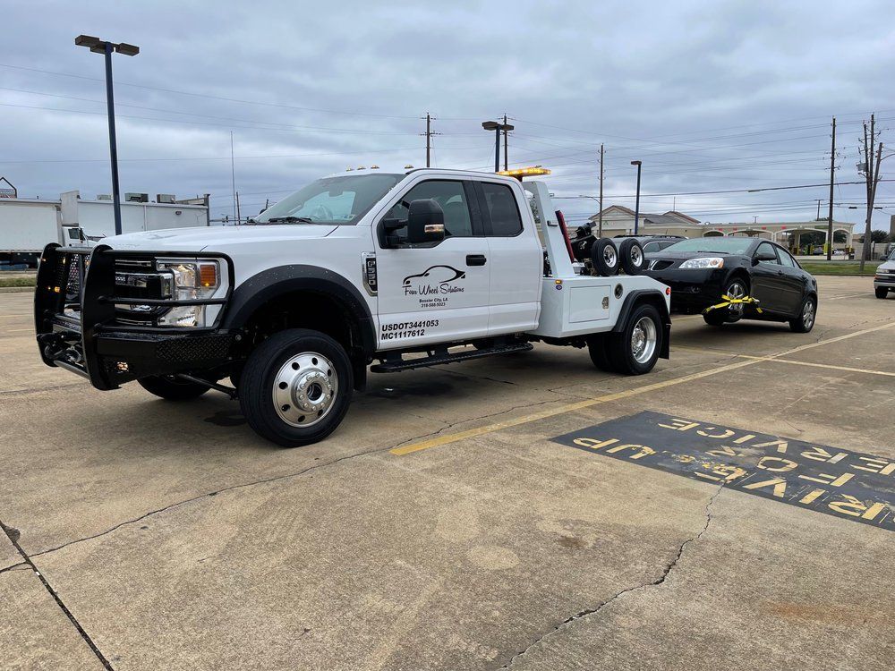 A white tow truck is towing a black car in a parking lot.