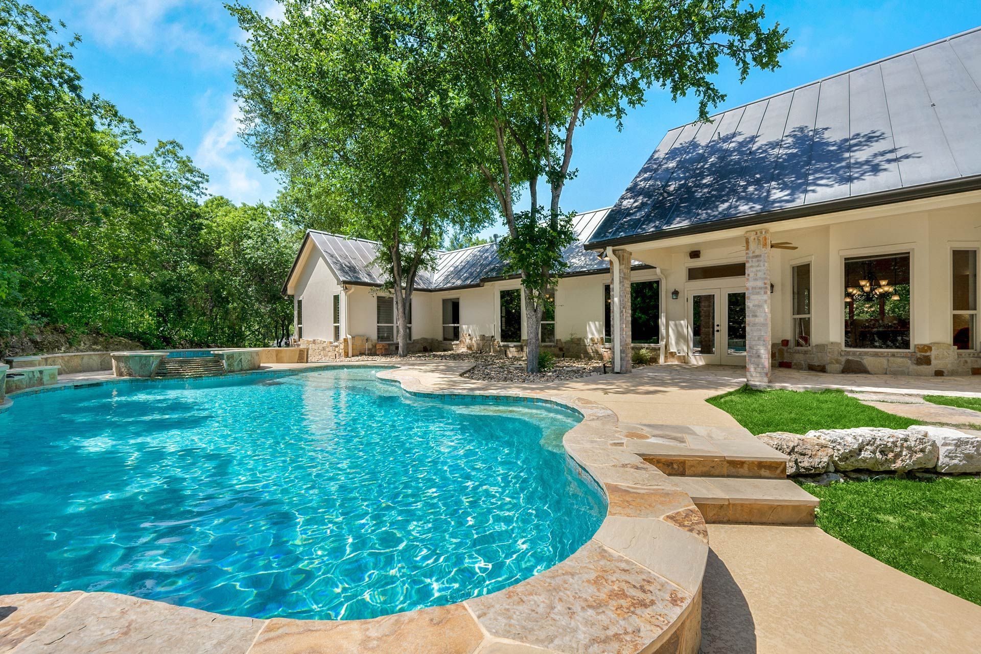Curved backyard pool beside stone house with porches, trees, and sunlight.