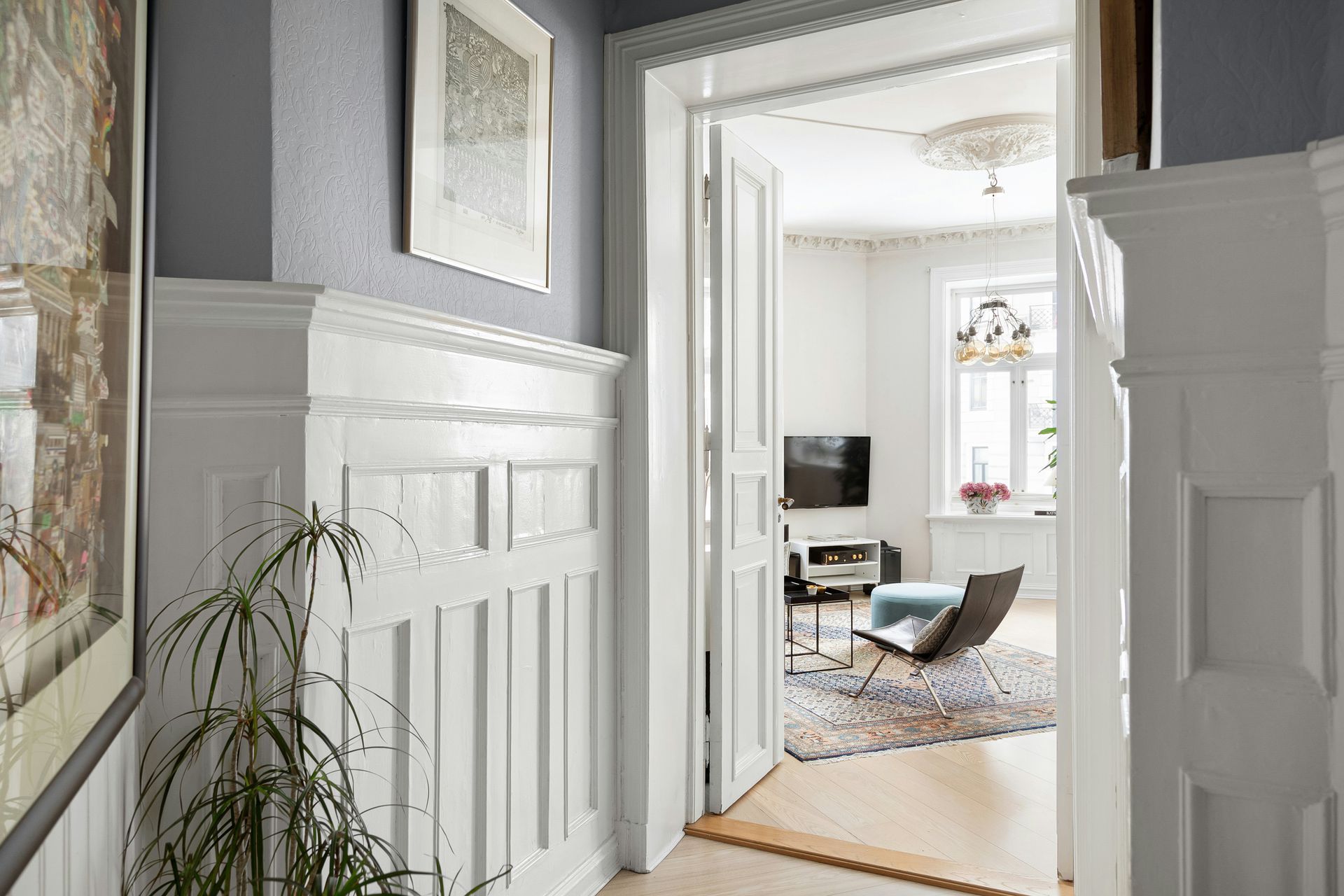 Bright hallway with white paneled walls leading into a sunlit room with a chair and rug