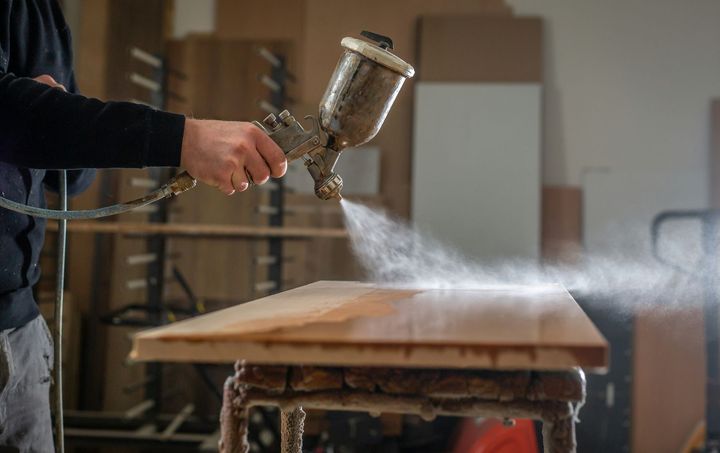 Person spraying white paint onto a wooden board with a spray gun in a workshop