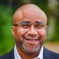 Man with glasses and beard smiles outdoors, wearing a blazer.