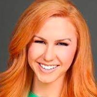 Woman with bright orange hair smiles at the camera, against a grey background.