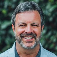 Smiling man with graying hair and beard, outdoors, blurred green background.