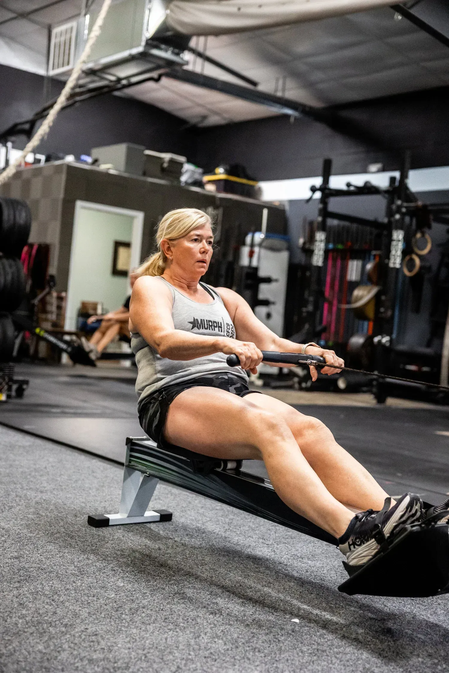 Woman rowing on a machine at a gym, pulling a handle towards her chest. She wears athletic clothing.