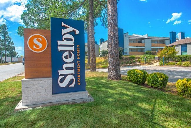 Sign for Shelby Apartment Homes with blue, orange, and white lettering on a lawn, with a building in the background.