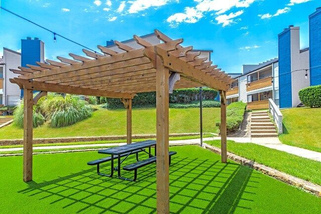 Pergola with picnic table on green lawn, blue sky. Buildings in background.
