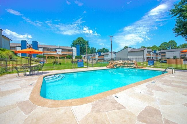 Swimming pool with clear blue water, patio furniture, and apartment buildings under a sunny sky.