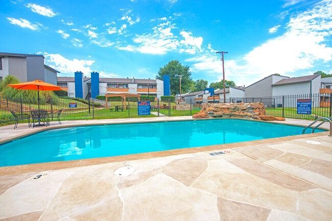 Swimming pool with a rock waterfall, surrounded by apartments, blue sky.