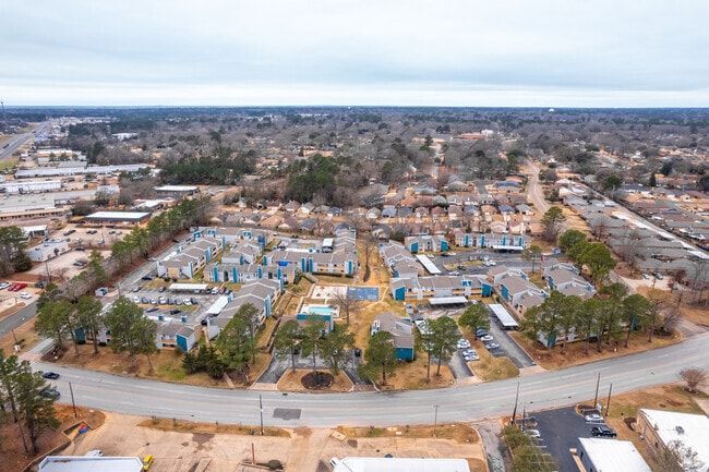 Aerial view of an apartment complex with blue roofs, surrounded by trees and roads. Overcast sky.
