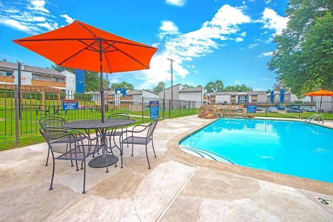 An orange umbrella shades a table and chairs by a swimming pool on a sunny day.