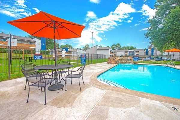 An orange umbrella shades a table and chairs by a swimming pool on a sunny day.