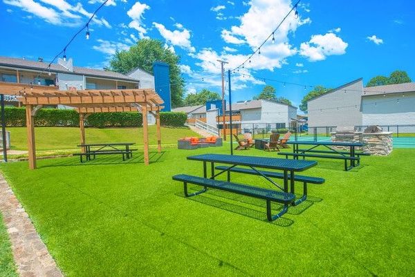 Outdoor green space with picnic tables, a pergola, and string lights on a sunny day.