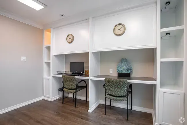 Two built-in desks with chairs, clock and computer in a white office space with wood flooring.