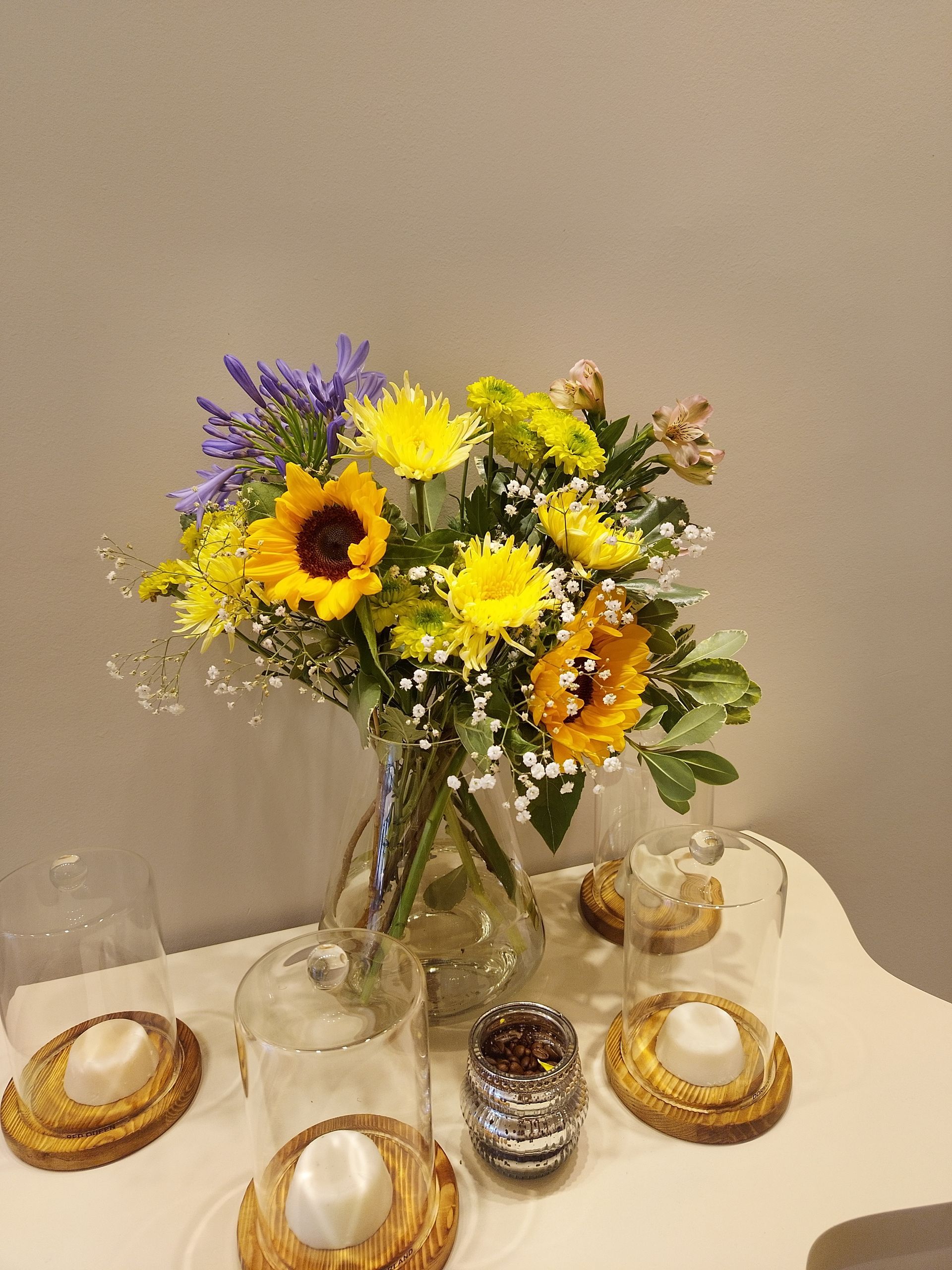 Ramo de girasoles amarillos, margaritas y flores violetas en un jarrón sobre una mesa blanca con velas.