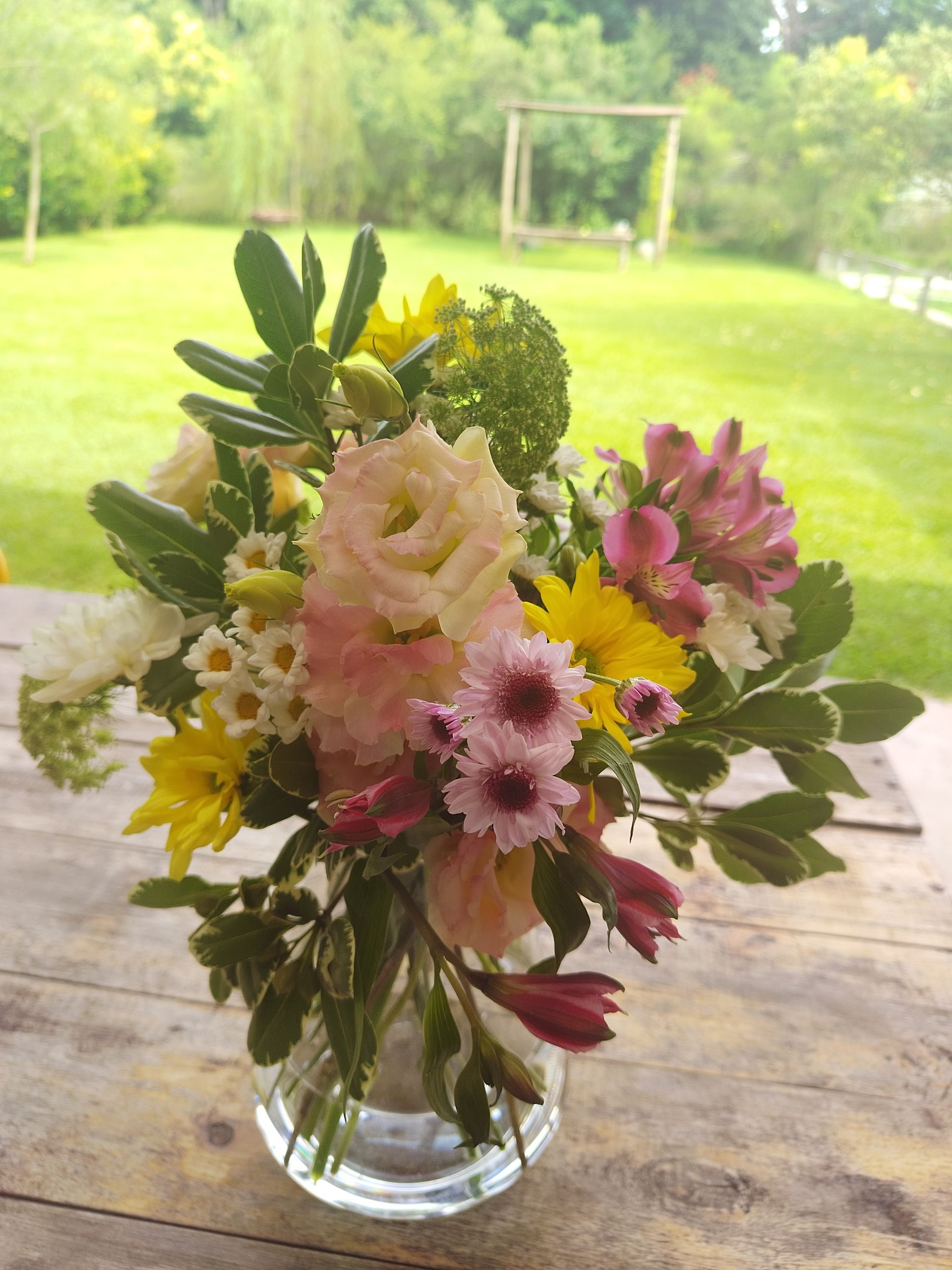Un ramo de flores de colores en un jarrón de cristal sobre una mesa de madera, con un fondo verde borroso.