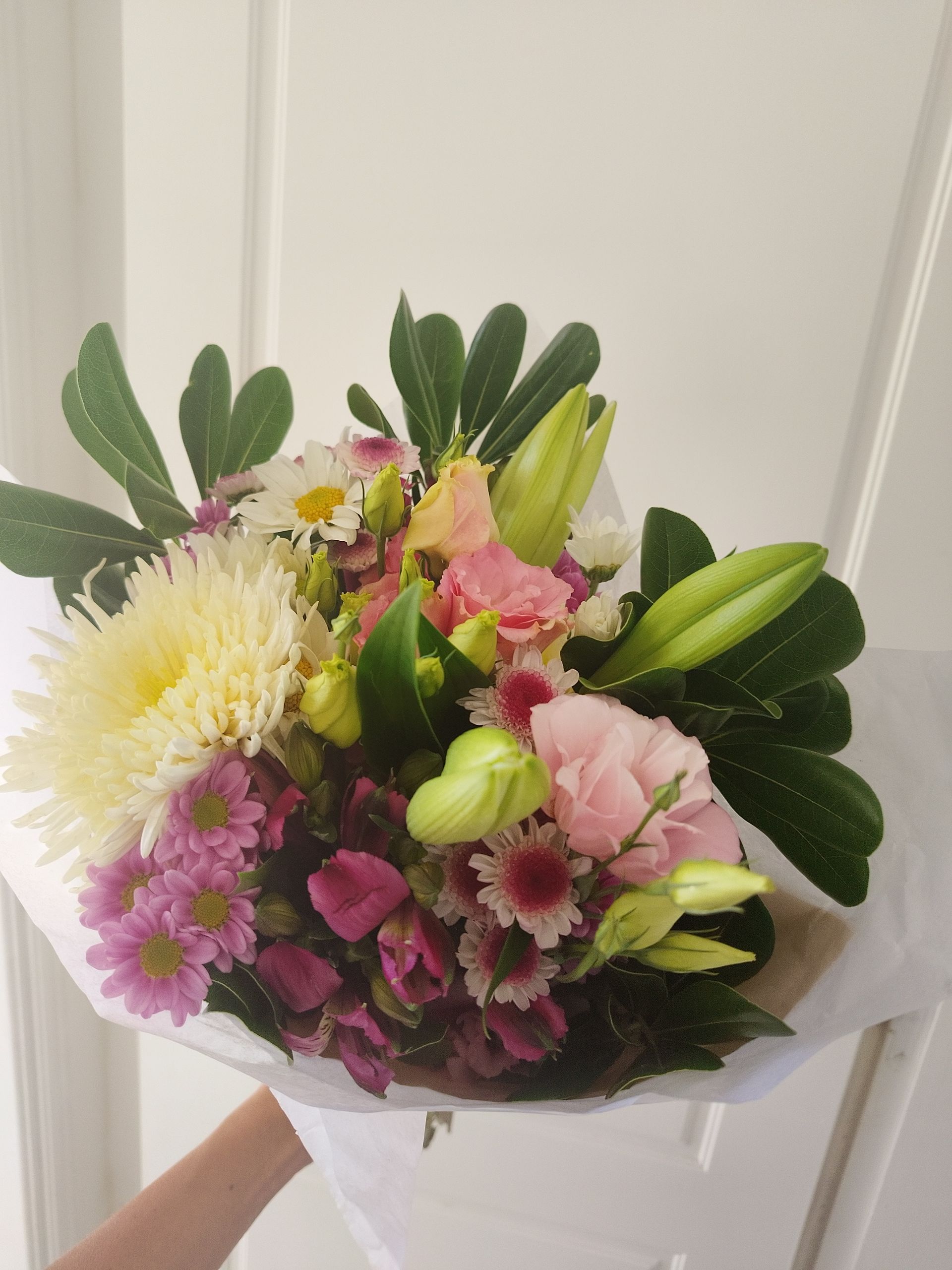 Ramo de flores de colores, envuelto en papel blanco, sostenido por una persona frente a una puerta blanca.