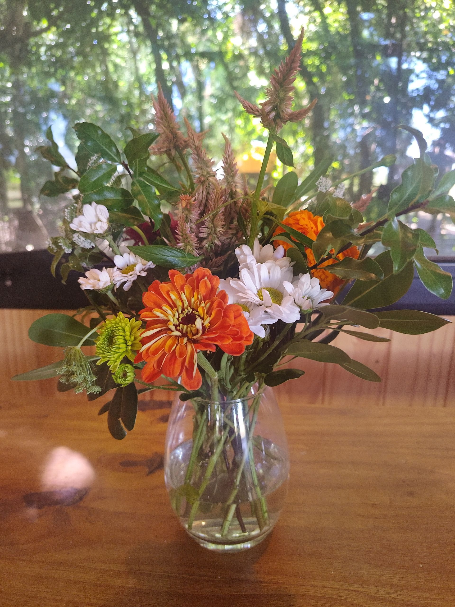 Ramo de flores naranjas y blancas en un jarrón de cristal sobre una mesa de madera, fondo verde borroso.