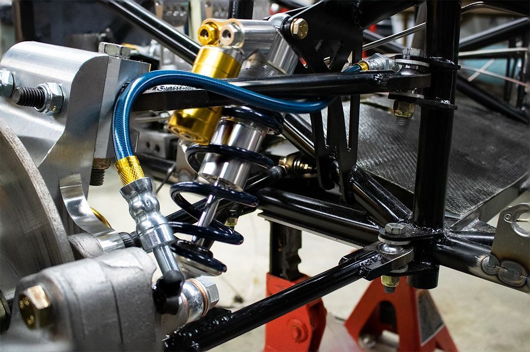 Close-up of a race car's suspension and shock absorber system. Blue brake hose, black frame, gold shocks.