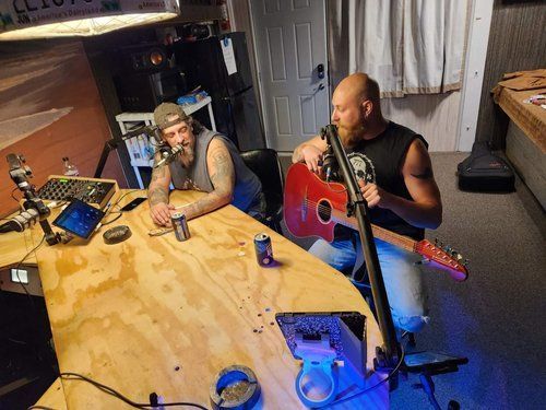 Two men are sitting at a table playing guitars and singing into microphones.