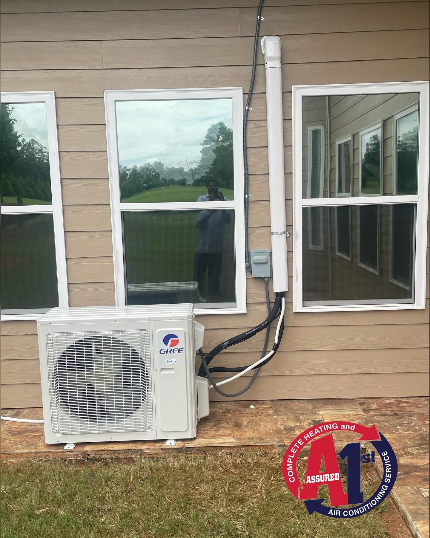 White air conditioning unit installed outside a house with windows. Piping and conduit run up the wall.
